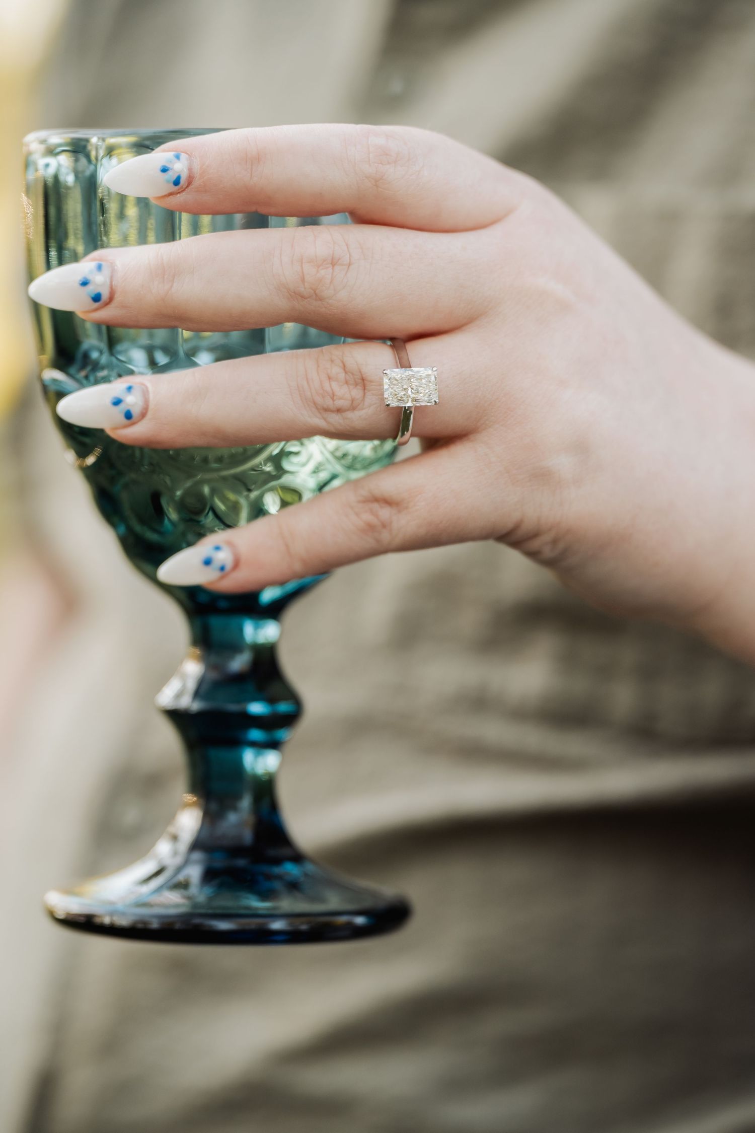 A hand holding a blue-tinted vintage glass goblet showcases floral nail art and a sparkling diamond engagement ring.