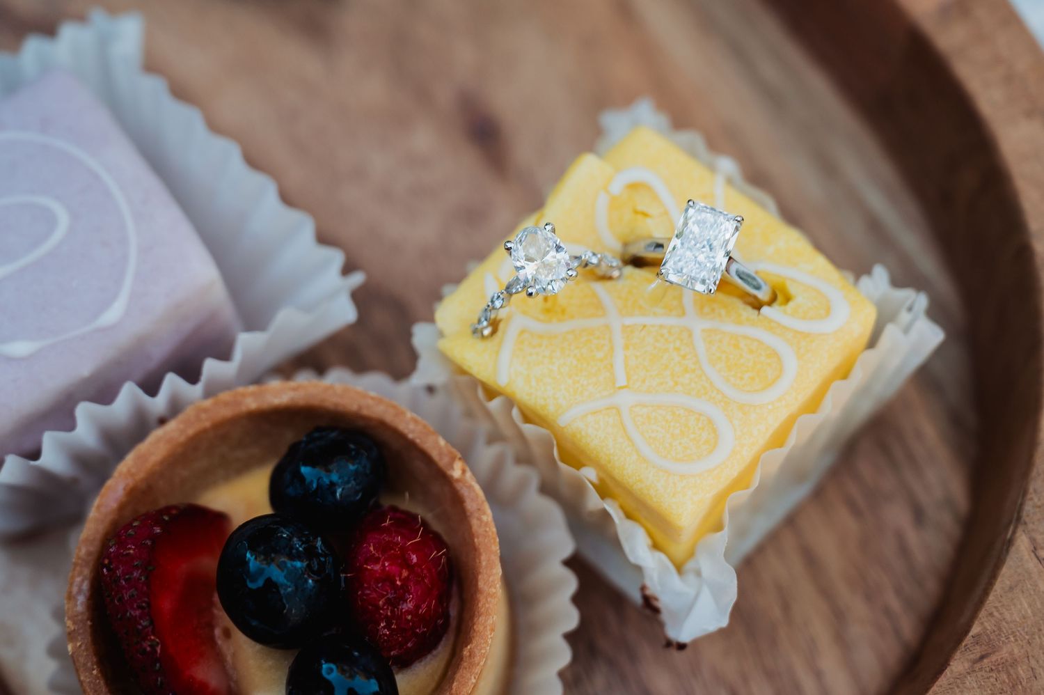 Wedding rings rest on a yellow dessert beside a fruit tart with berries on a wooden surface.