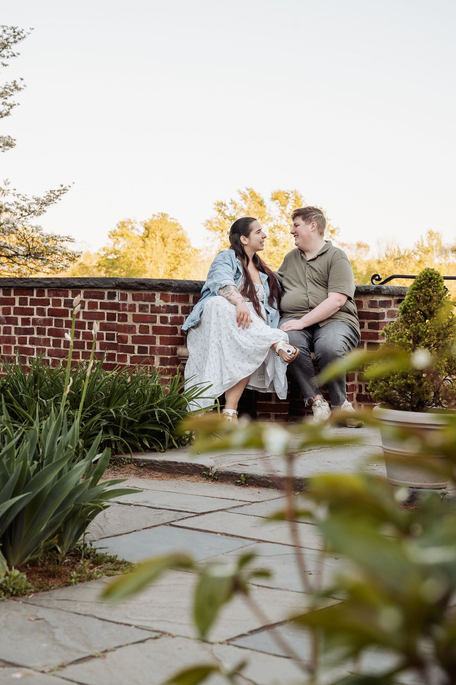 A couple sits together on a brick wall surrounded by garden landscaping during golden hour.