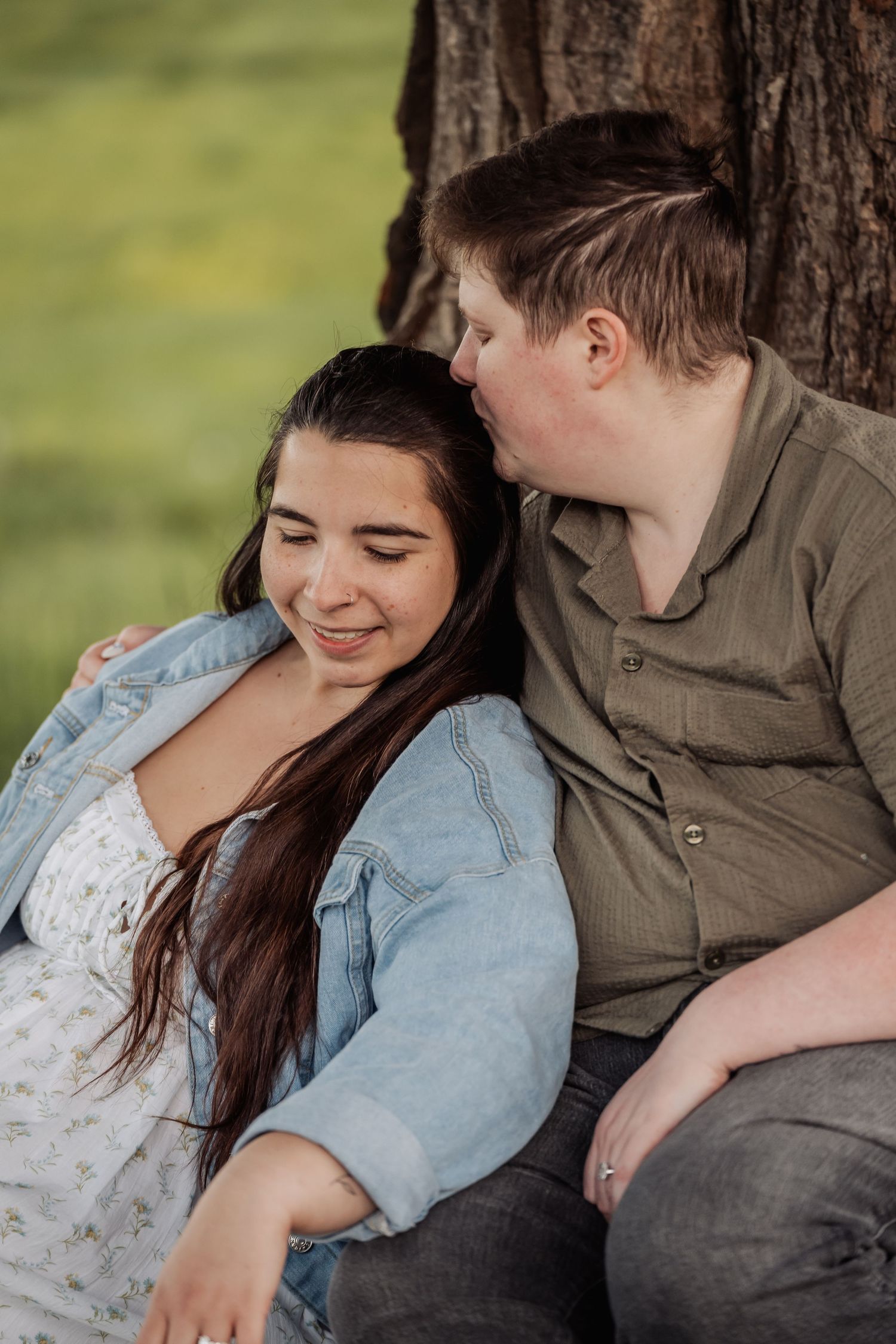 A tender moment shared between a couple sitting against a tree trunk in casual denim and olive clothing.