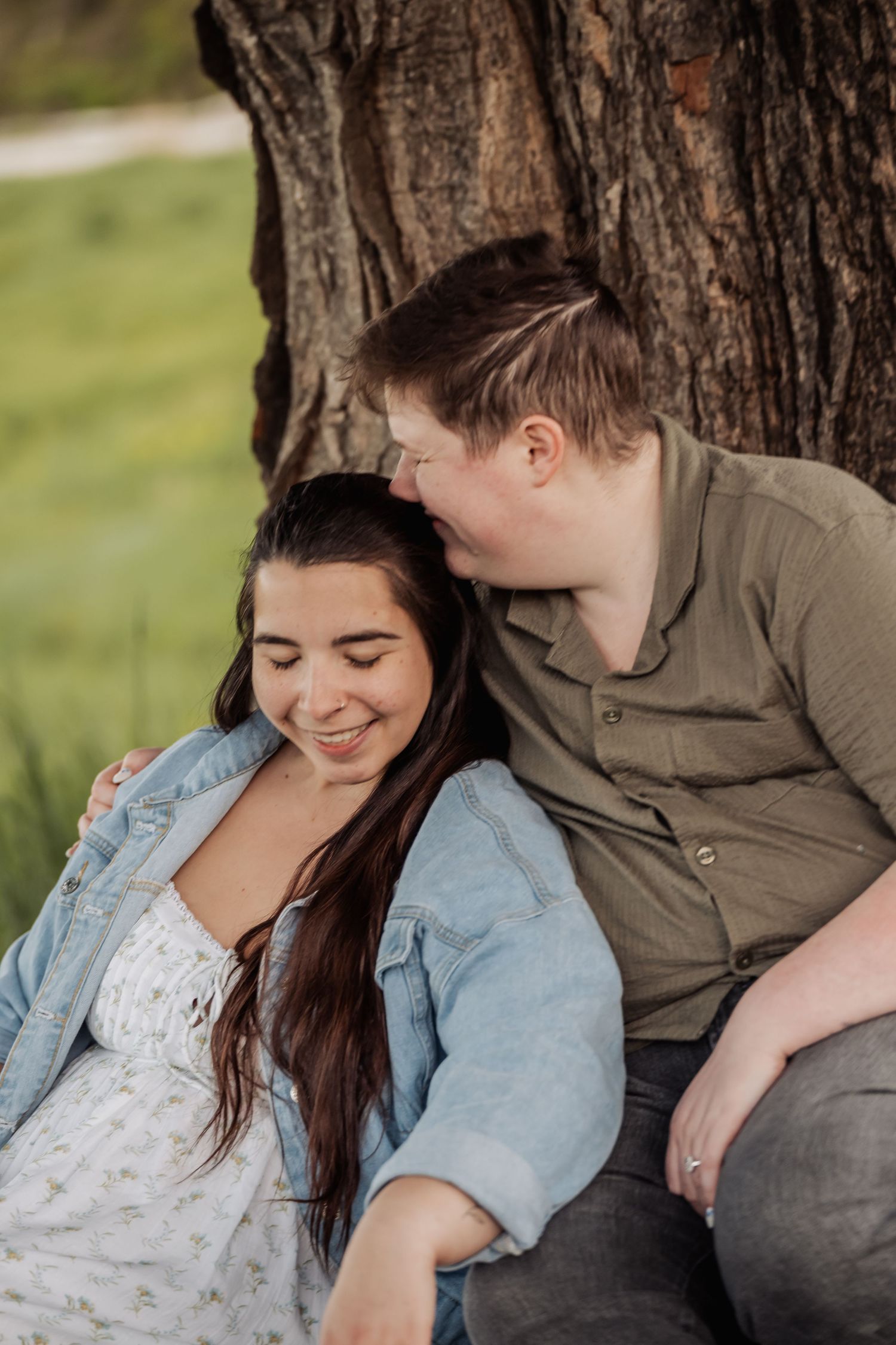 A heartwarming couple sits together beneath a large tree trunk, sharing an intimate moment outdoors.