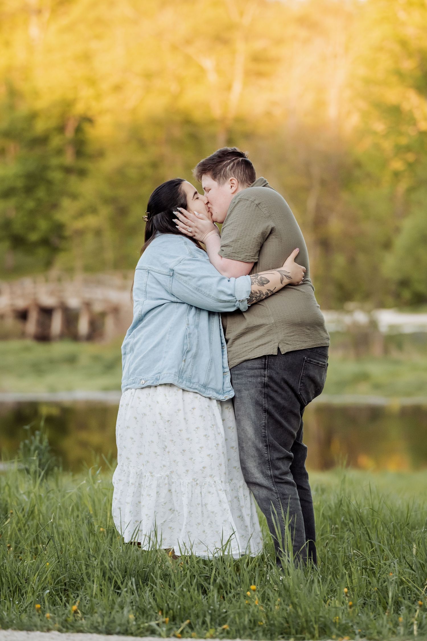 A couple shares a romantic kiss by a lake with autumn foliage in the background during a golden sunset.