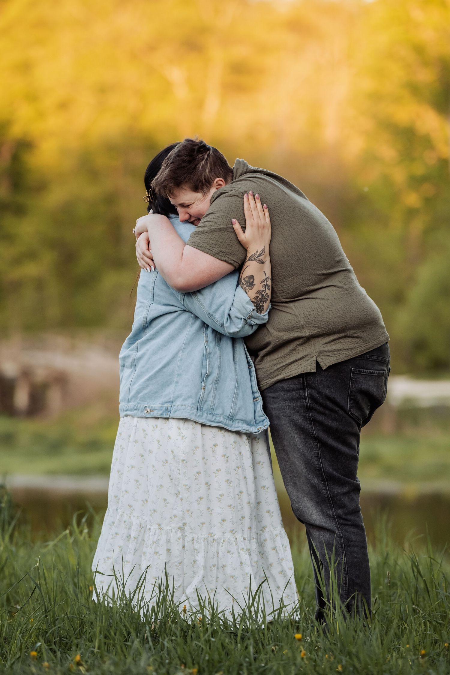 A couple embrace tenderly in a meadow at sunset with golden autumn foliage in the background.