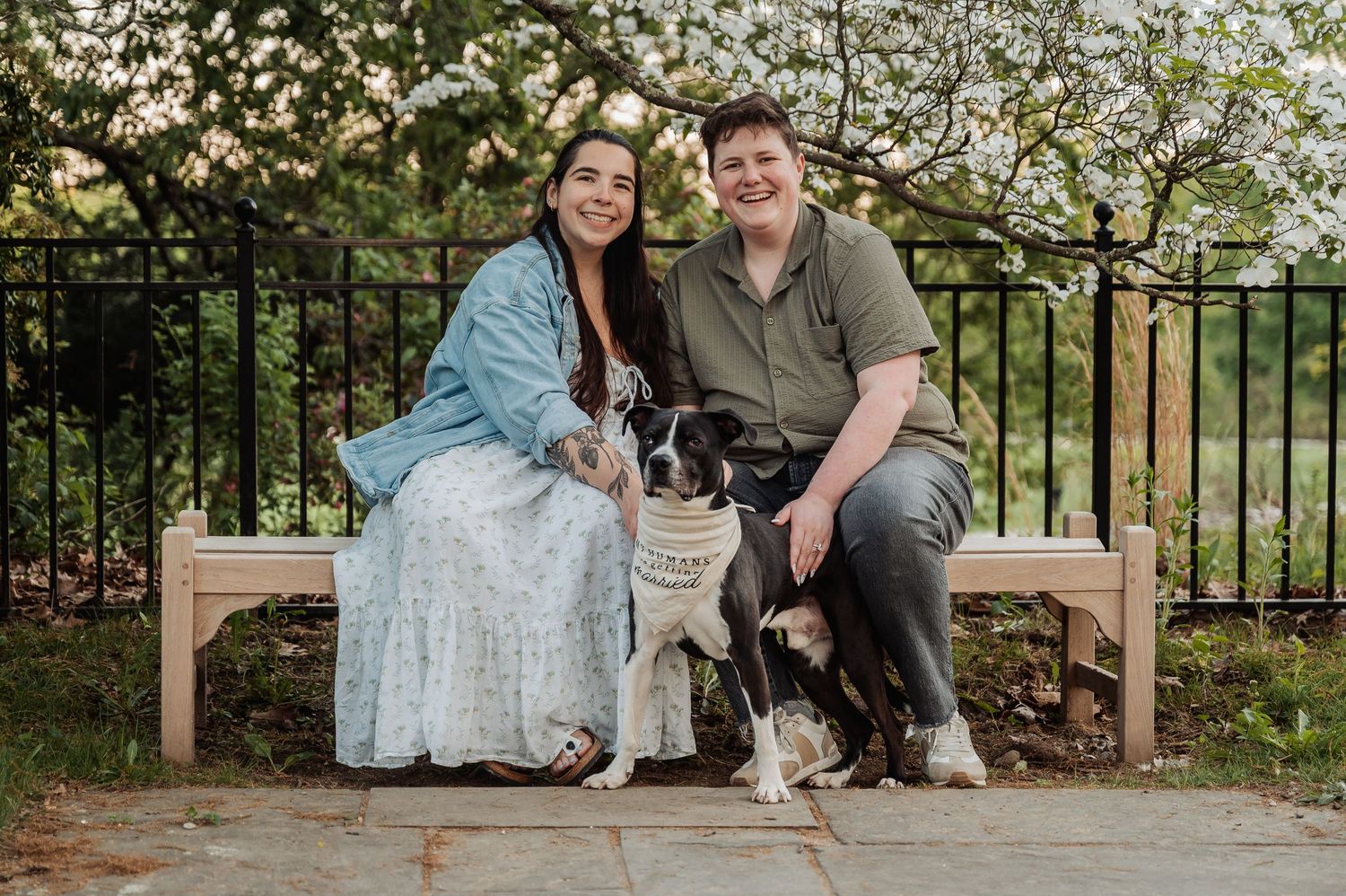 A couple sits on a wooden bench with their small dog under blooming spring trees.