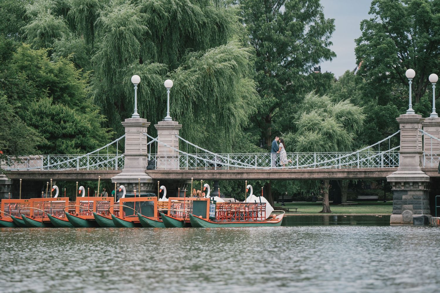 A historic footbridge with ornate pillars spans across a lake with orange boats visible below. Boston Engagement & Wedding Photographer