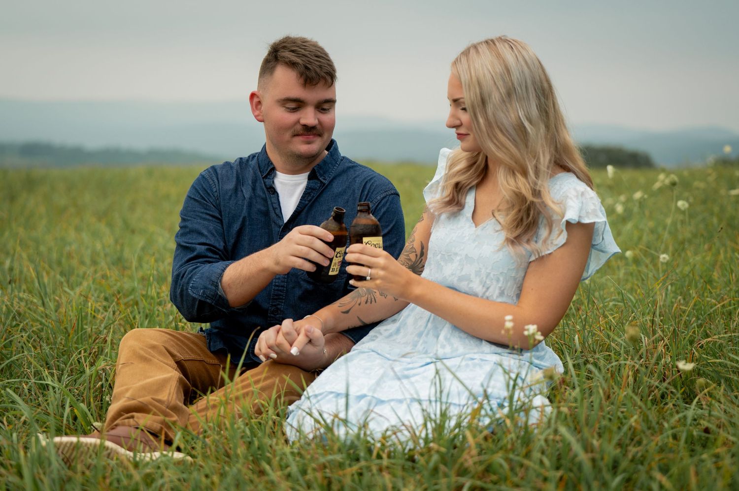 A couple sits together in a blooming meadow holding drinks and enjoying a sunny afternoon outdoors.