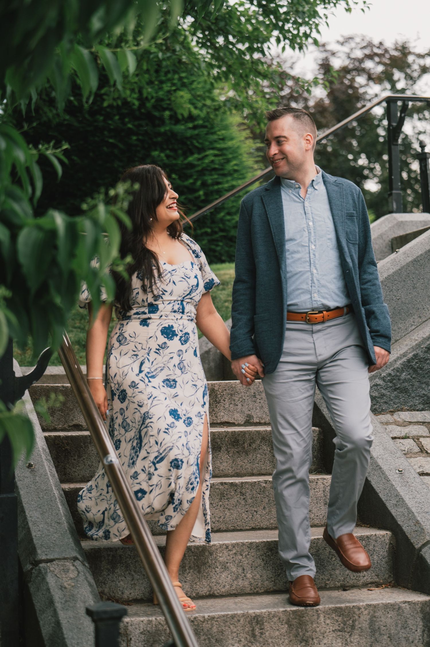 A well-dressed couple walks hand in hand down stone steps surrounded by green foliage. Boston Engagement & Wedding Photographer