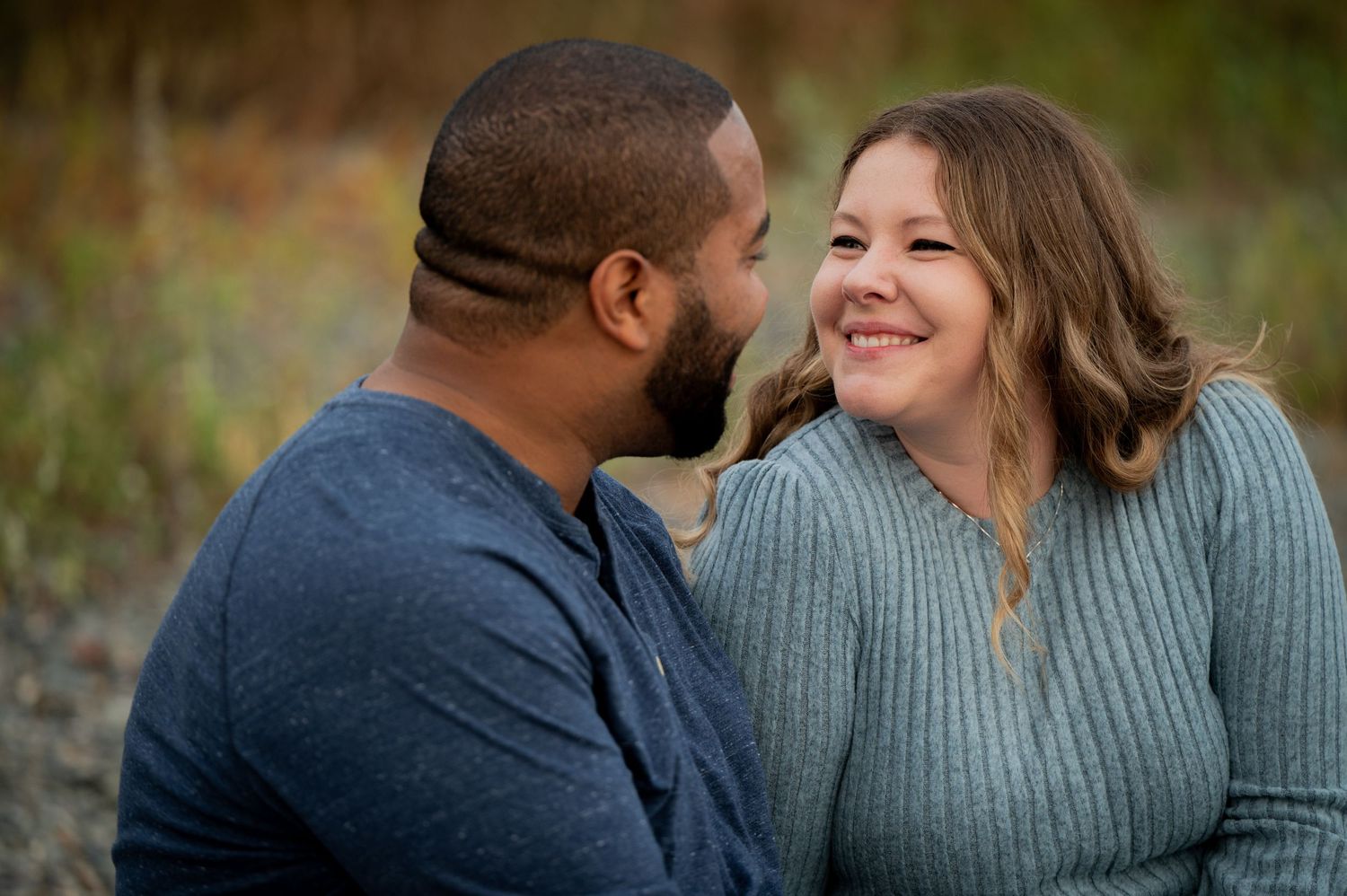 A couple shares a joyful moment together while sitting outdoors in casual sweaters during an autumn photoshoot.