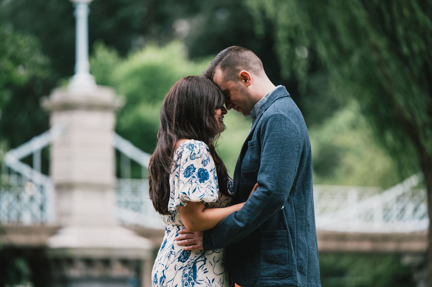A romantic couple shares an intimate moment in front of an ornate bridge in a lush garden setting. Boston Engagement & Wedding Photographe
