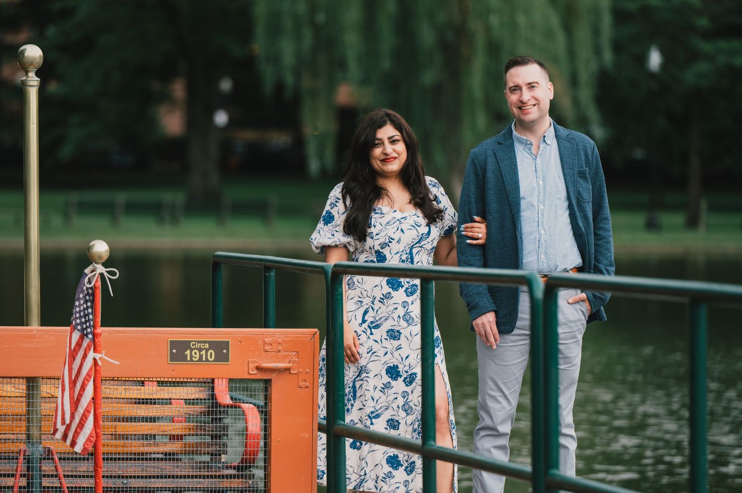 A couple standing on a dock with an American flag and boats in the background. Boston Engagement & Wedding Photographer