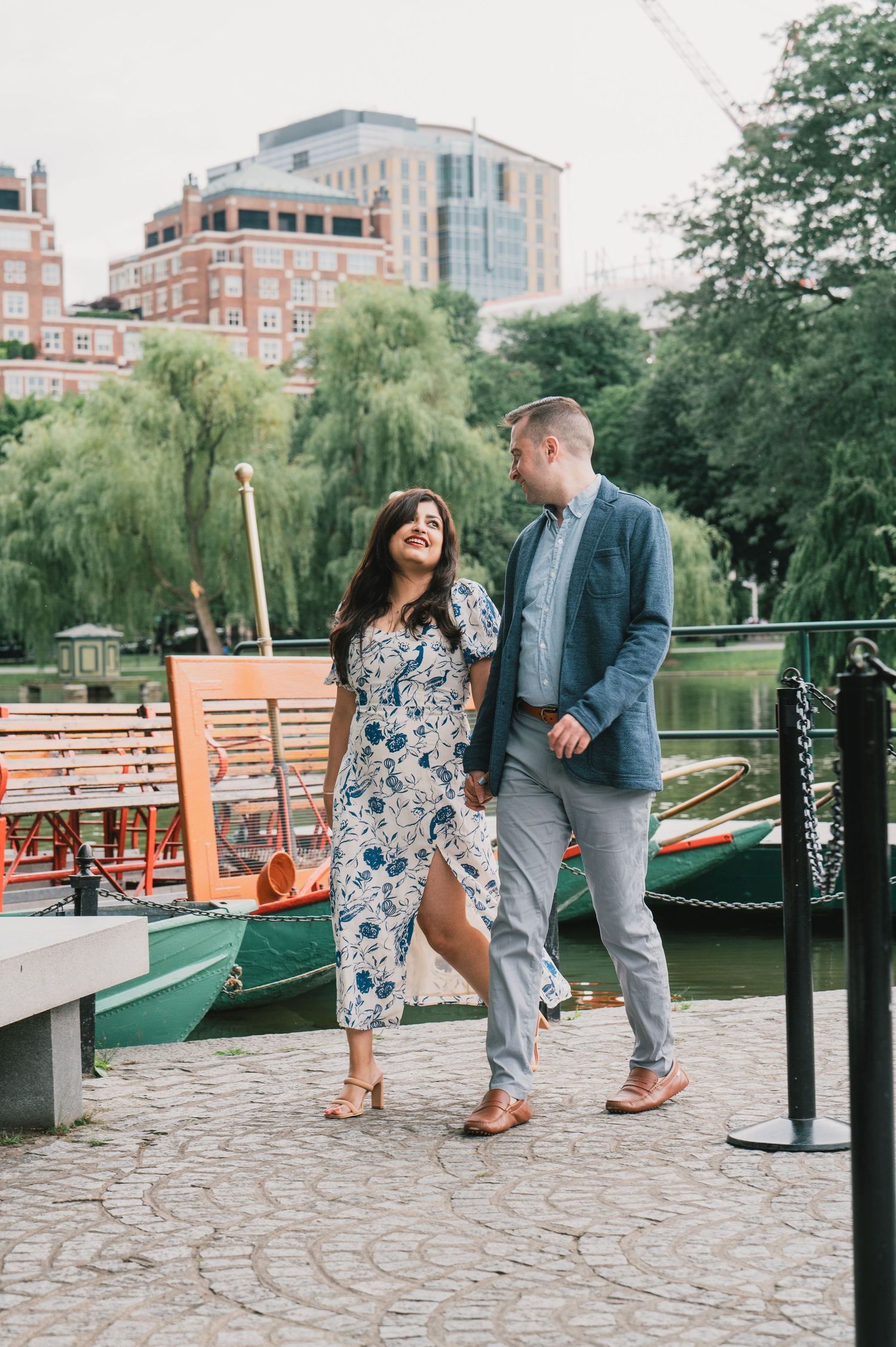 A couple walking along a waterfront path with boats and weeping willows in the background. Boston Engagement & Wedding Photographer