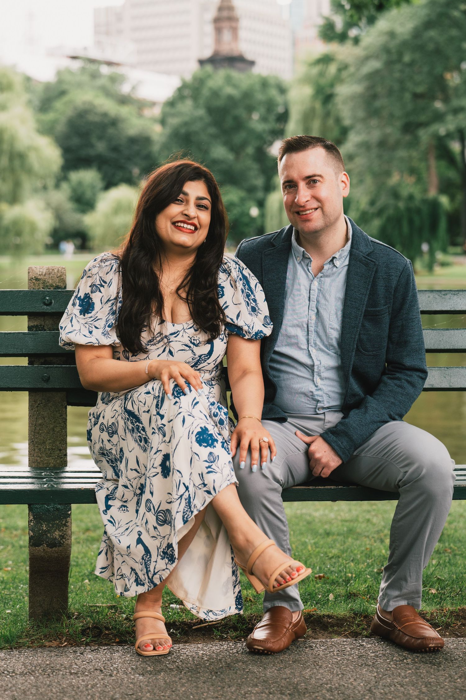 A couple sitting on a park bench smiling with city buildings and trees in the background. Boston Engagement & Wedding Photographer