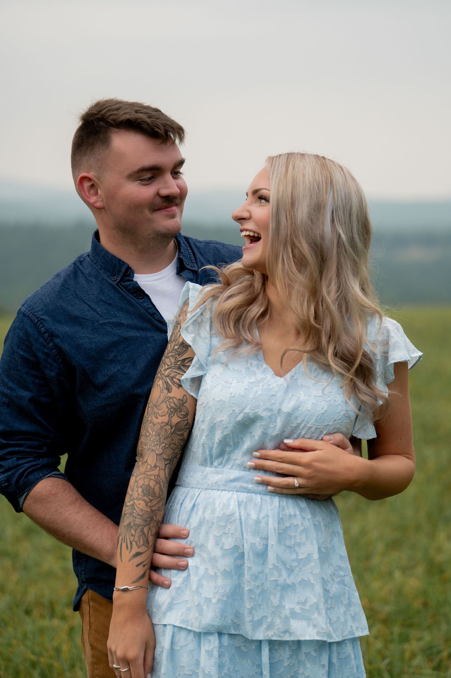 Happy couple embrace in a field at sunset as she laughs joyfully wearing a light blue dress while he shows off his tattooed arm.