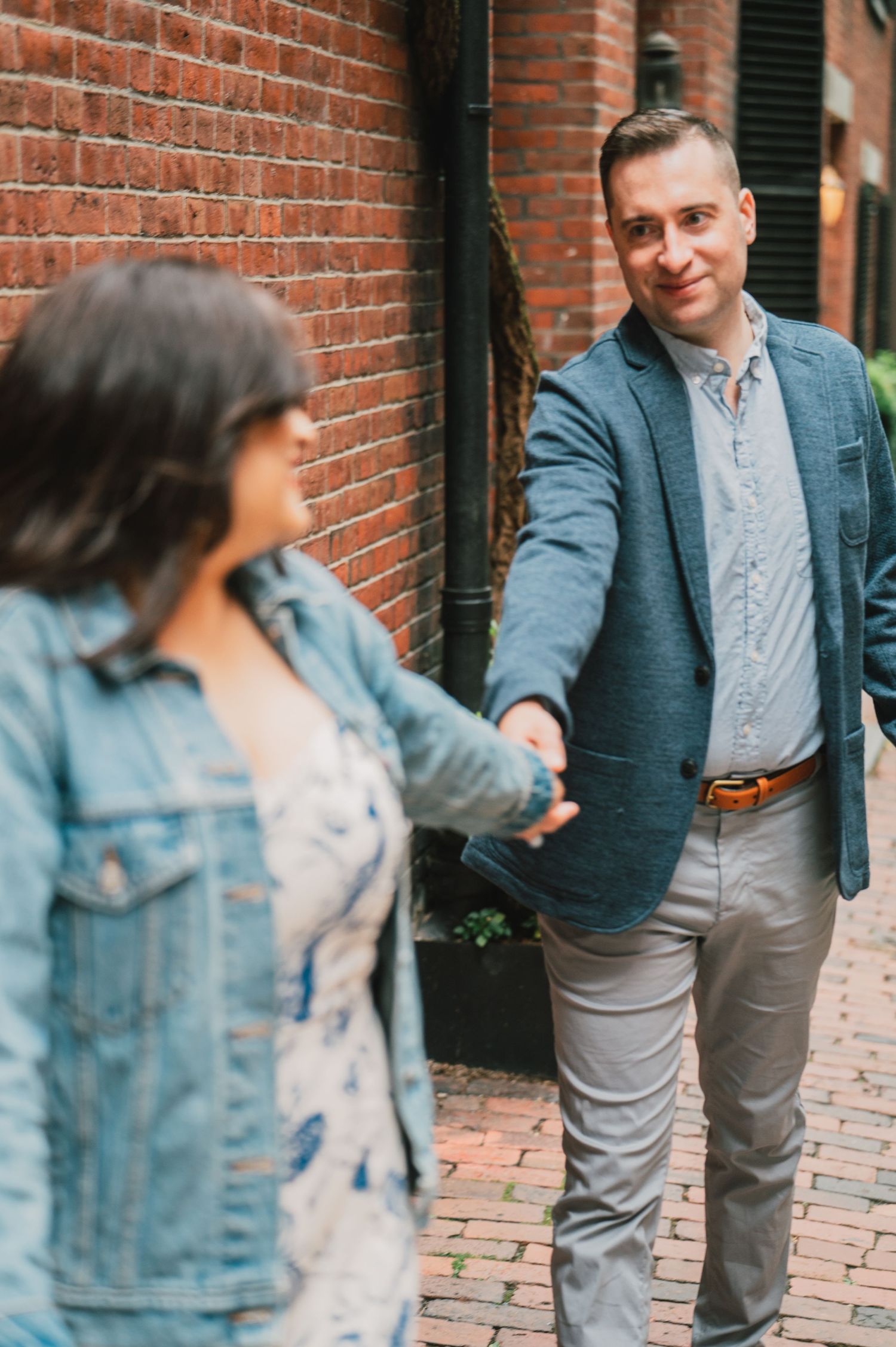 A couple holding hands and smiling while walking on a brick-paved street. Boston Engagement & Wedding Photographer
