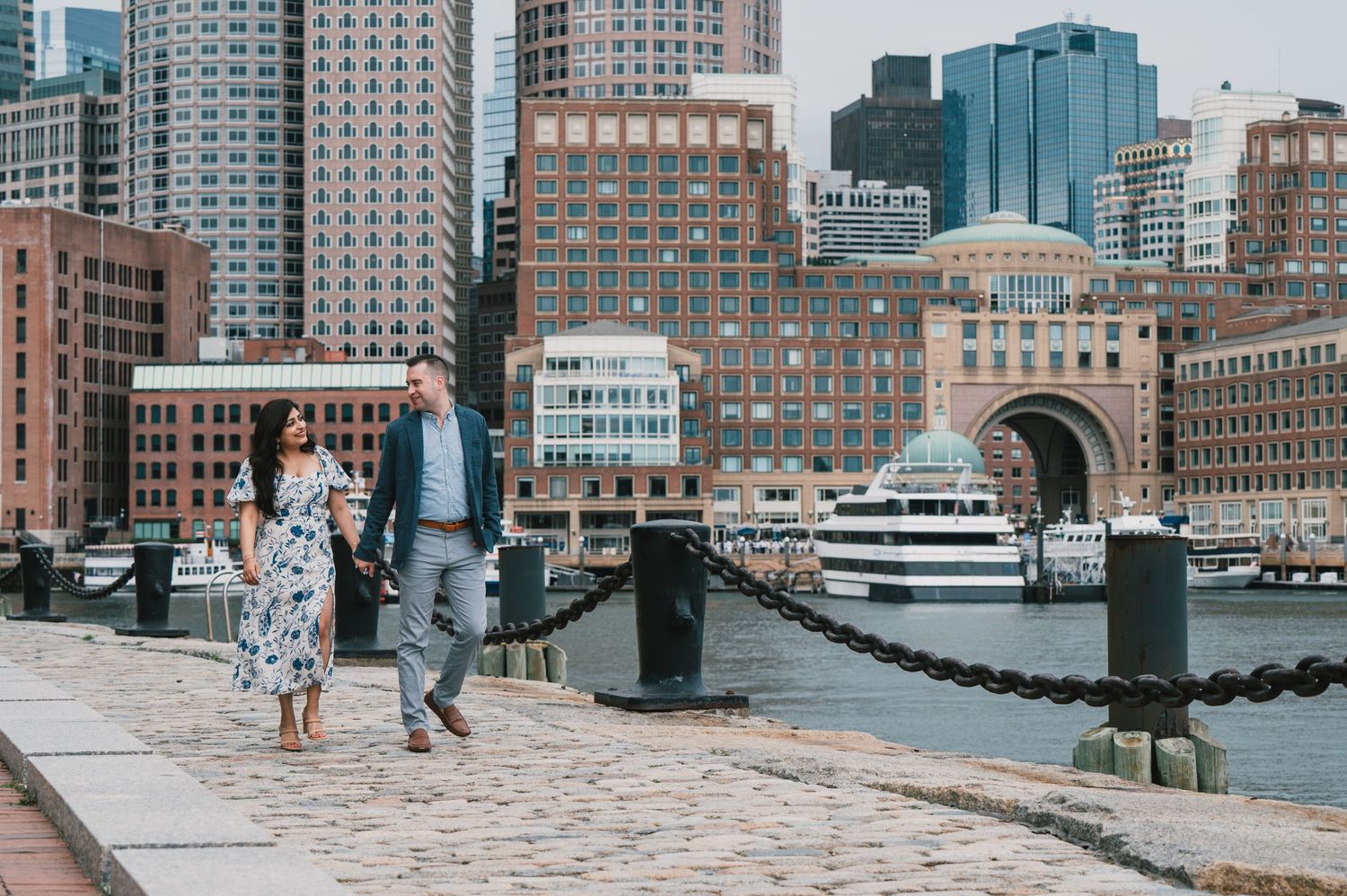 A couple strolls along the Boston Harbor waterfront with city skyscrapers in the background. Boston Engagement & Wedding Photographer