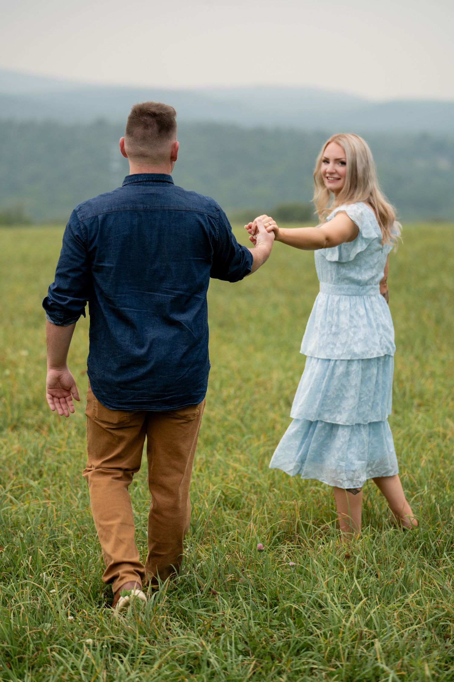 A couple dancing together in a grassy field with mountains in the background on a misty summer day.