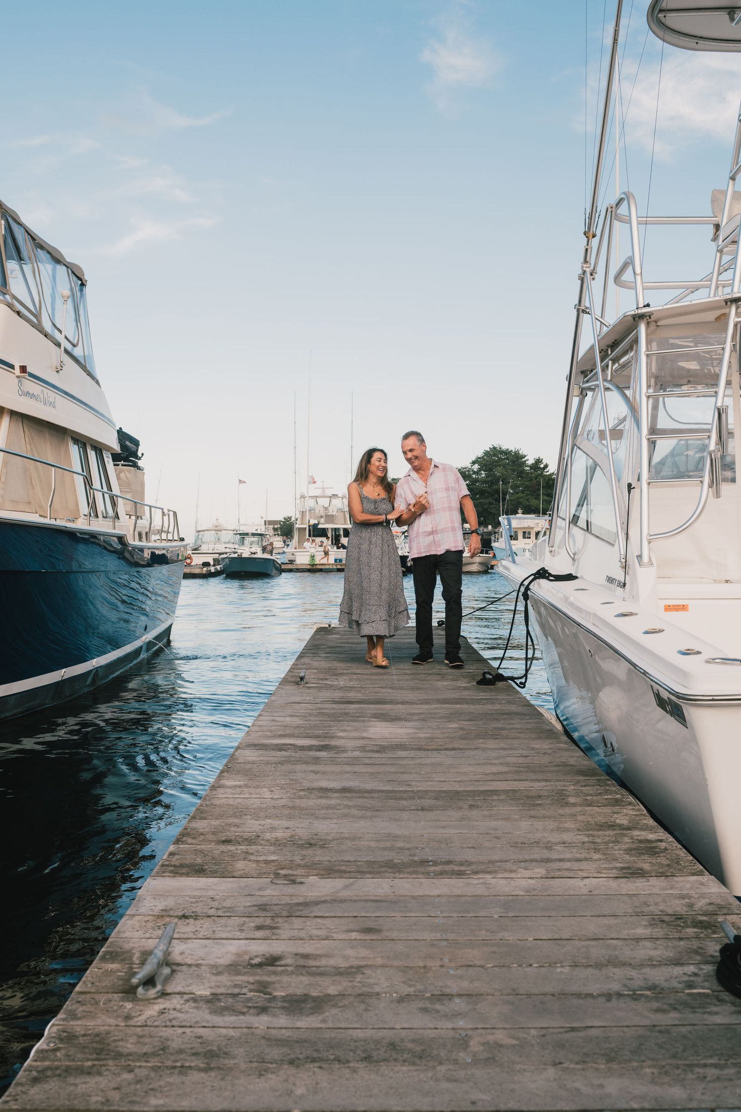 Two people stand together on a wooden dock surrounded by boats in a marina on a sunny summer day.