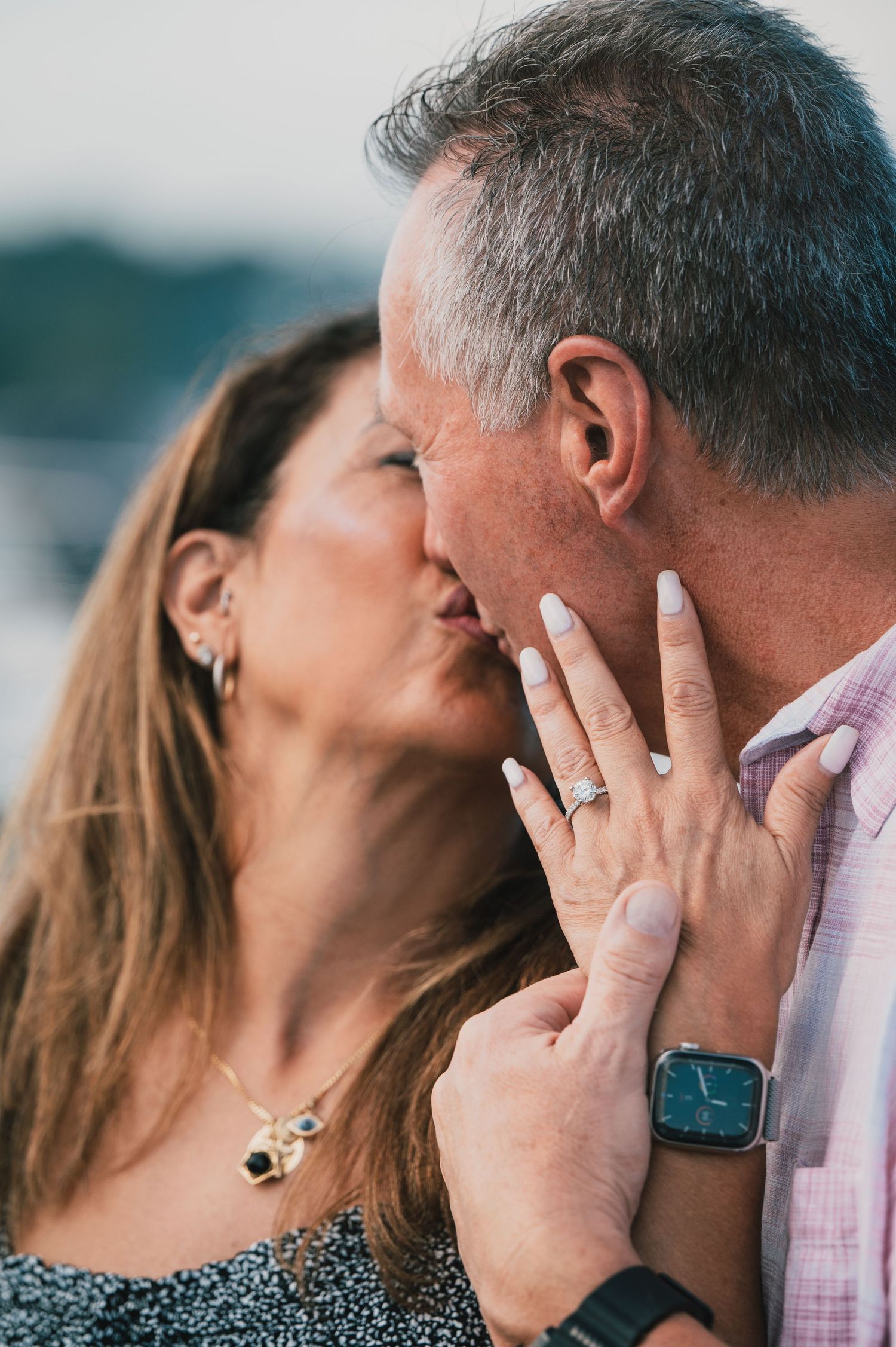 Close-up of a romantic mature couple sharing an intimate kiss outdoors with mountains in background and engagement ring visible.