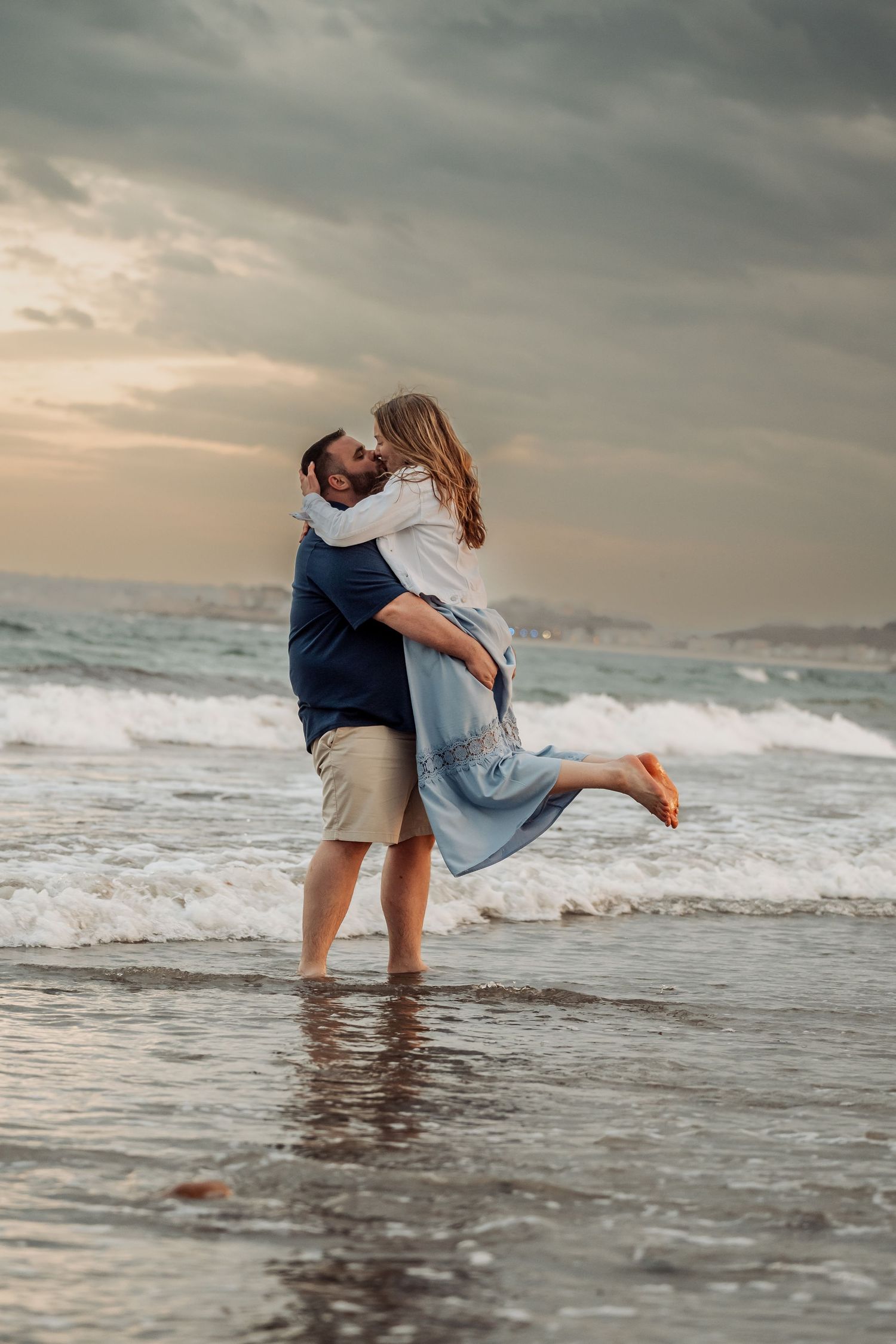 A romantic couple embraces on a misty beach at sunset as waves crash behind them against a moody sky.