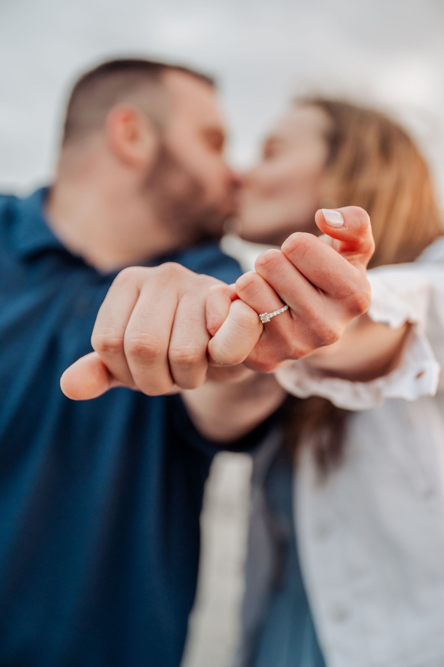 Close-up of intertwined hands showing engagement ring as couple shares intimate moment in background.