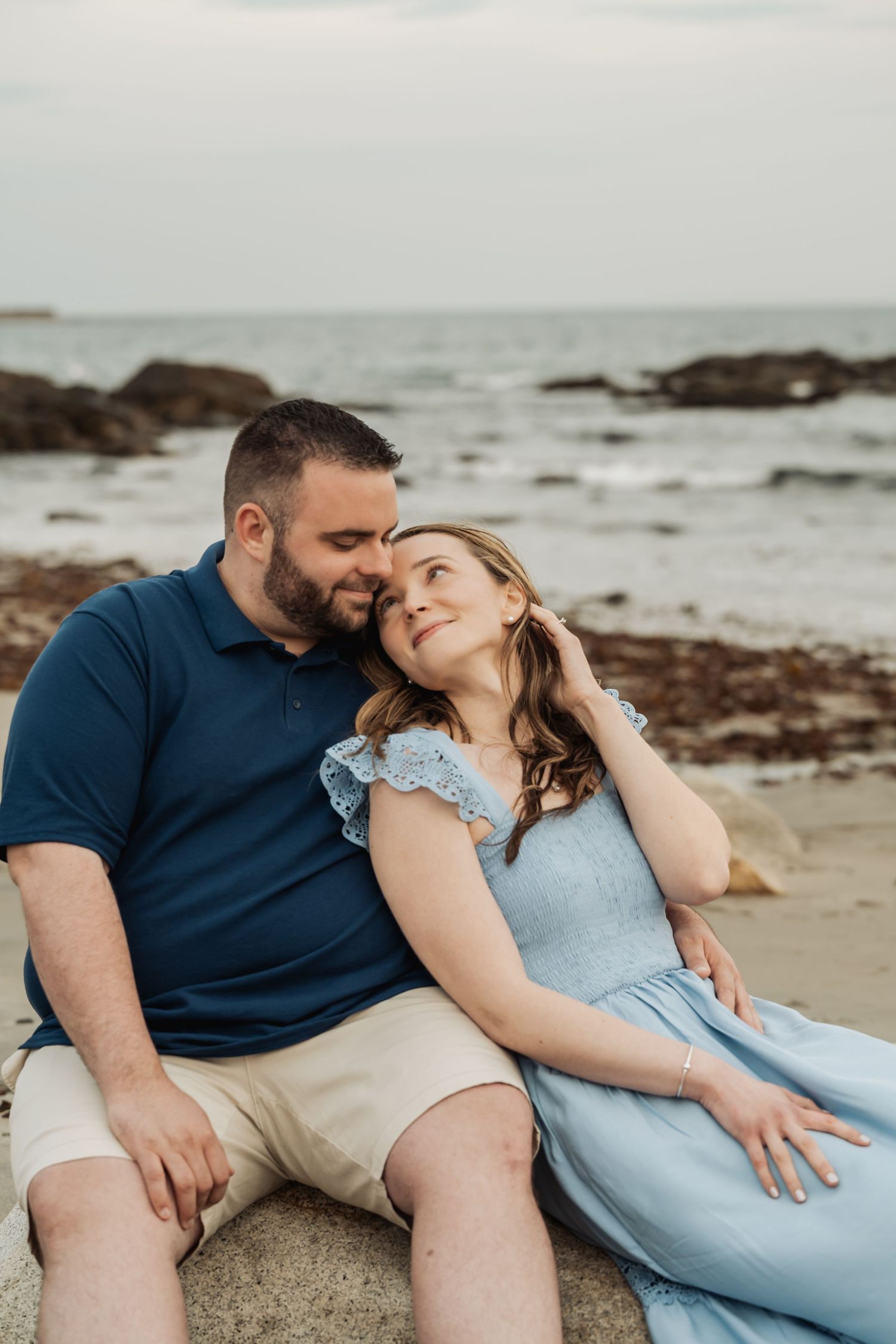 Couple shares intimate moment while sitting on rocky beach during sunset beside the ocean.