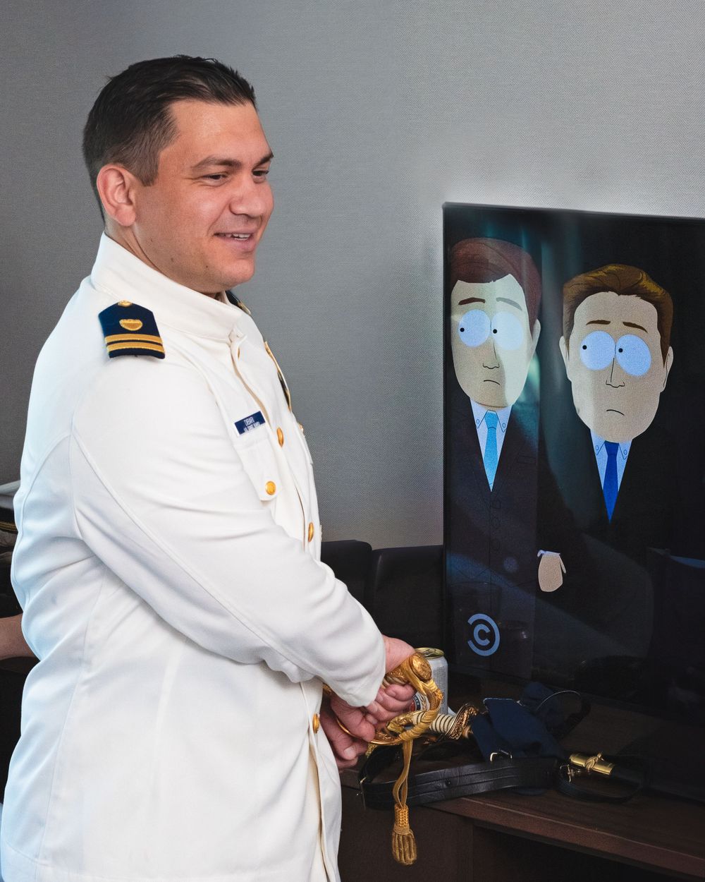 A naval officer in dress whites stands near a TV displaying an animated show.