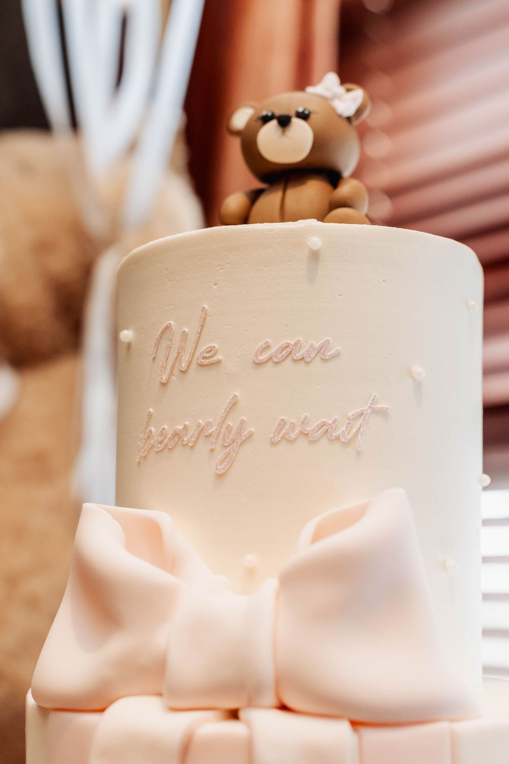 White tiered cake with pink bow and teddy bear topper featuring script lettering.