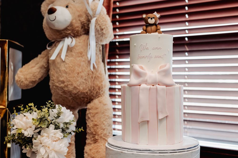 Elegant white baby shower cake with pink bow and teddy bear decorations beside floral arrangements.