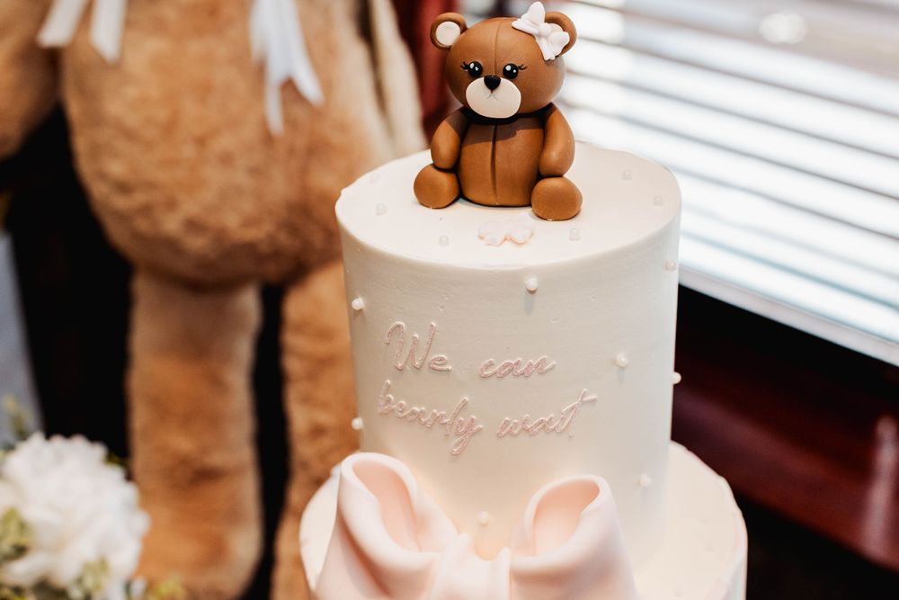 White tiered cake with pink bow and cute teddy bear topper for baby celebration.