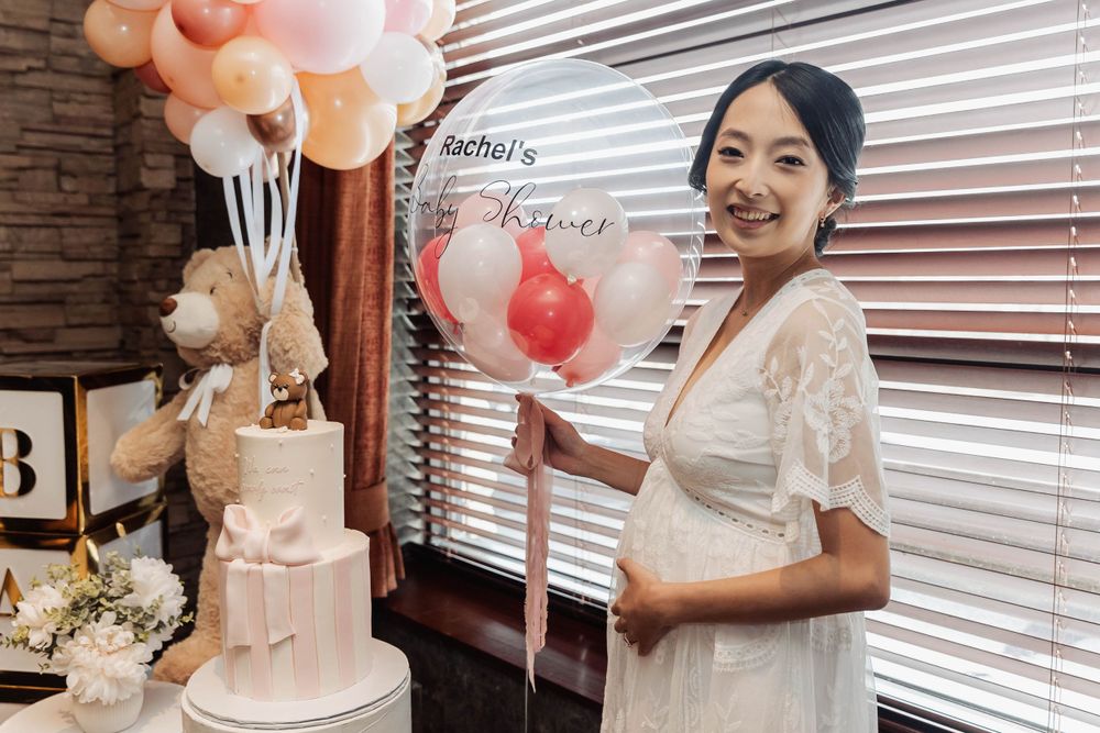 A smiling figure in a white lace dress poses with a pink balloon and tiered cake at a baby shower celebration.