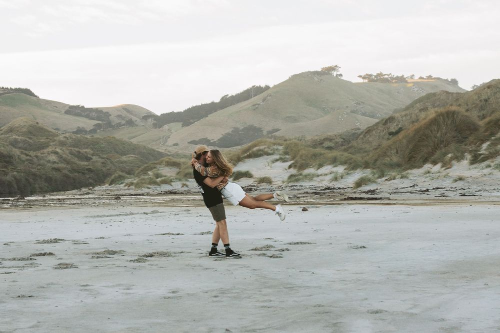 A couple embraces on a scenic beach with rolling hills in the background at sunset.