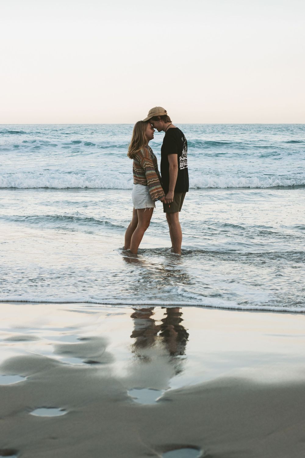 A romantic couple stands in shallow ocean waves at sunset with their reflection visible in the sand.