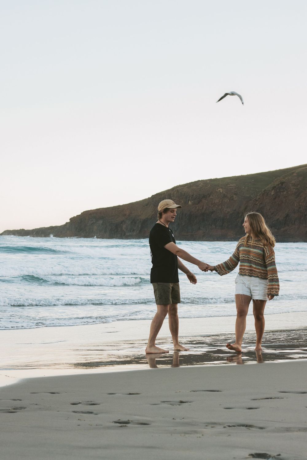 A couple holds hands while walking along a scenic coastal beach with cliffs and a seabird overhead.