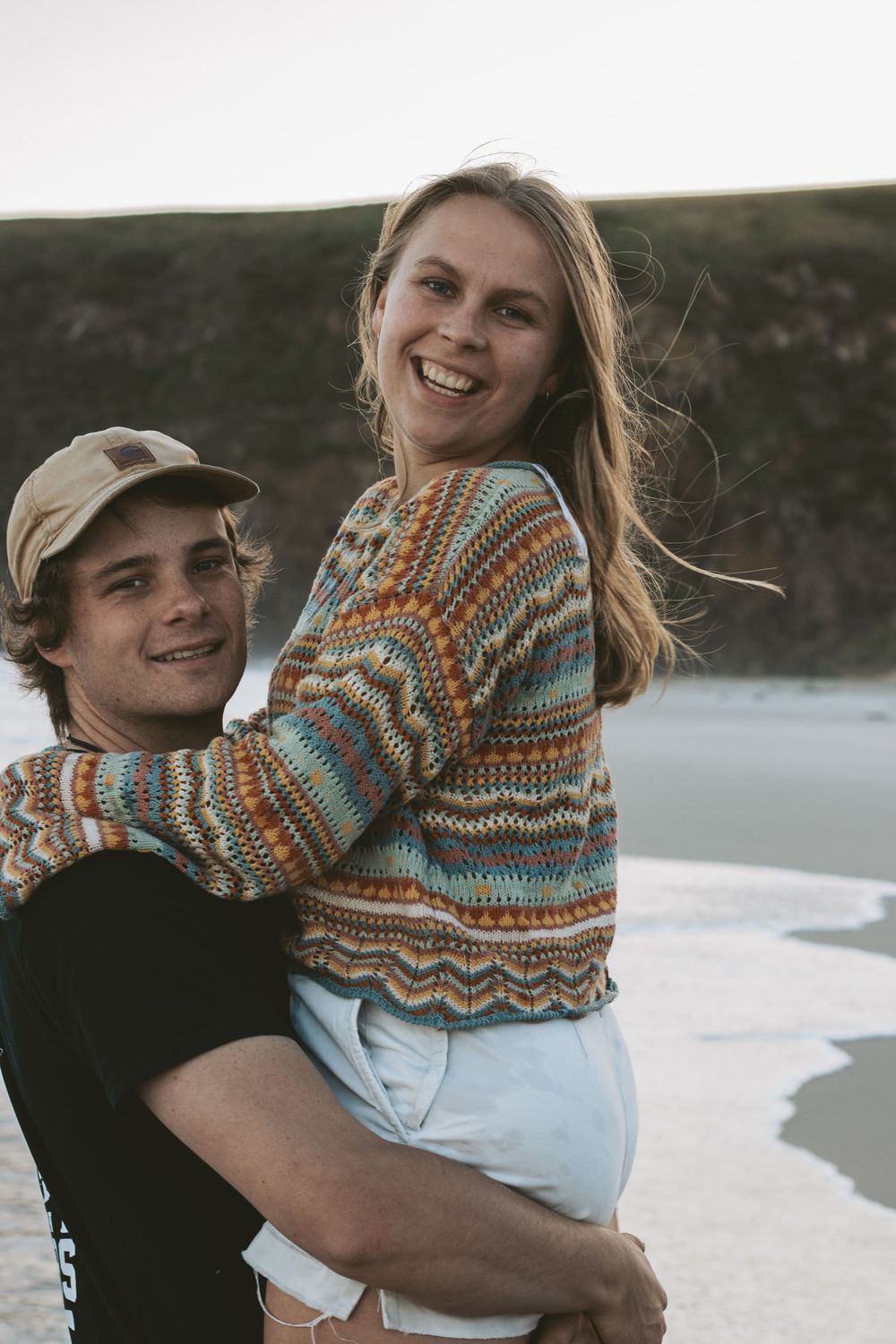 A couple in casual beachwear embraces and smiles against a coastal backdrop.