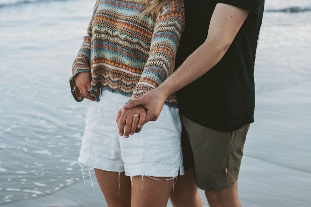 Close-up of a couple holding hands on the beach, wearing casual summer attire.