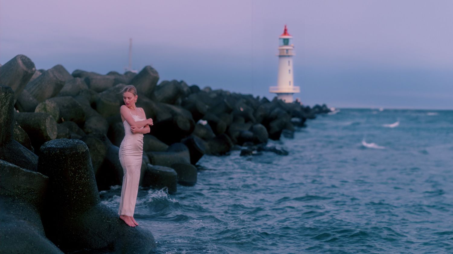 Barefoot bride in a long silky dress stands by the ocean with the lighthouse in the background