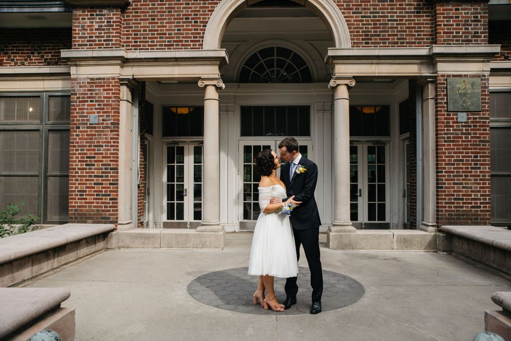 A wedding couple shares an intimate moment in front of a classic brick building with columns.
