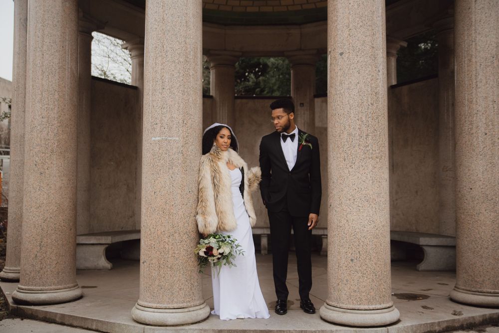 A wedding couple poses elegantly between classical stone columns in formal attire.