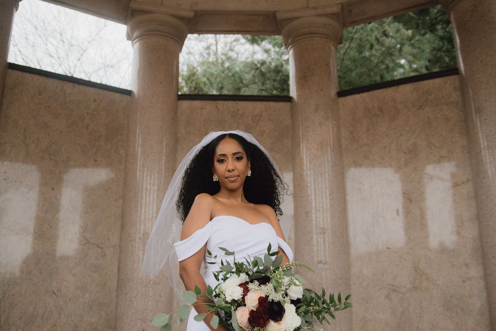 A bride in an off-shoulder white dress holds a bouquet of flowers while standing under a stone gazebo.