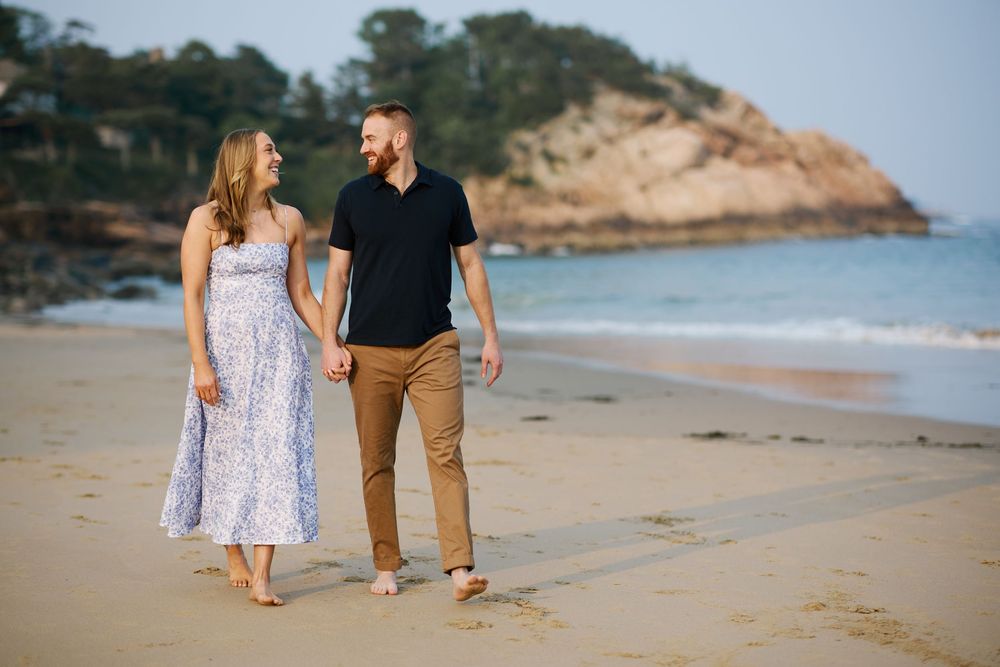 A couple walks hand in hand along a scenic sandy beach with cliffs and ocean waves visible in the distance.