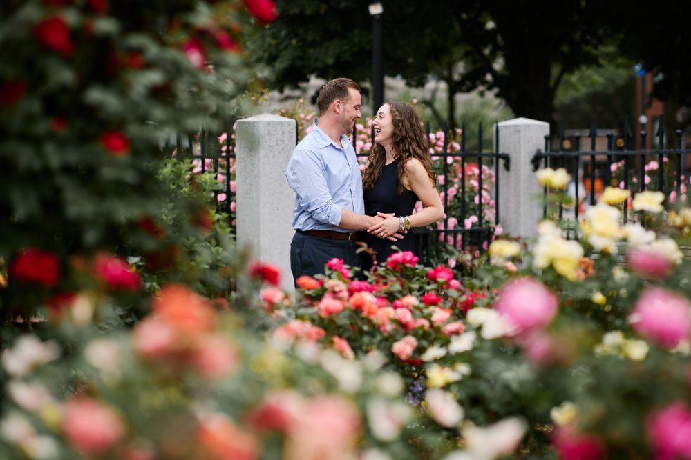 Two people embrace in a romantic moment surrounded by blooming rose bushes in a garden setting.
