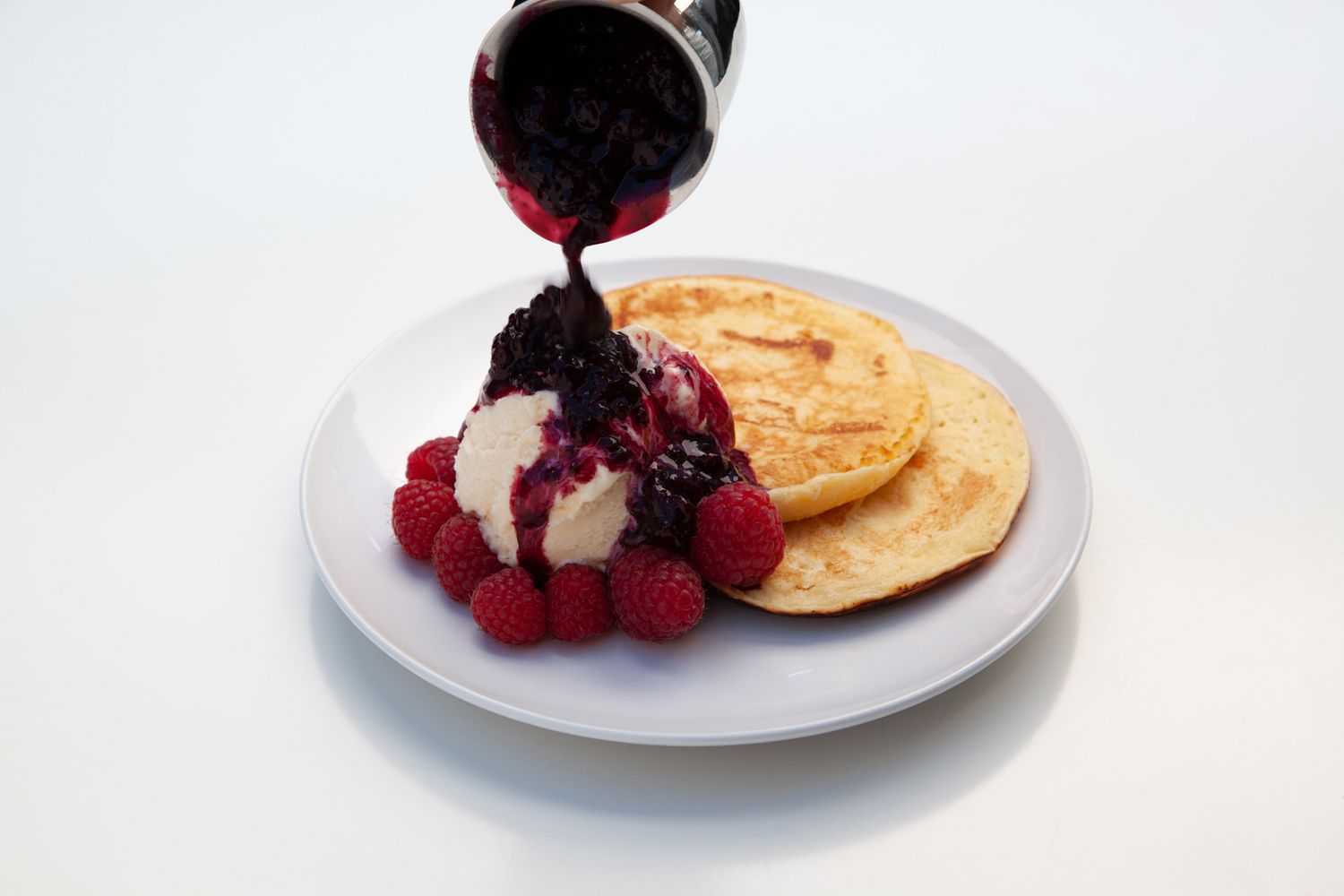 Red berry sauce being poured over ice cream and pancakes on white plate.