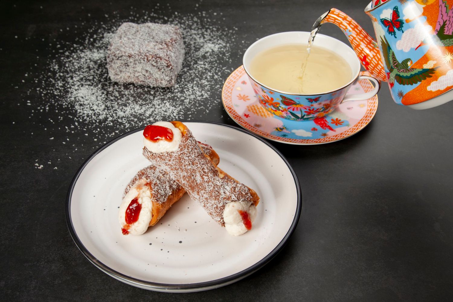 Cannoli with powdered sugar served alongside decorative teacup with hot beverage.