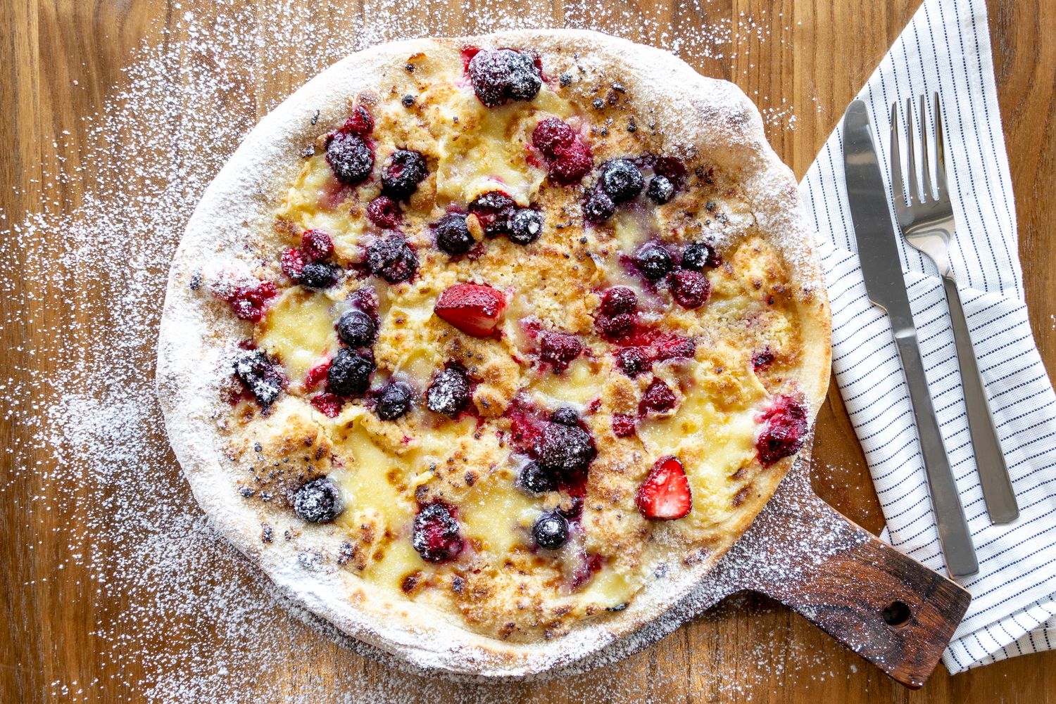 Homemade fruit pizza topped with fresh berries and powdered sugar on wooden board.