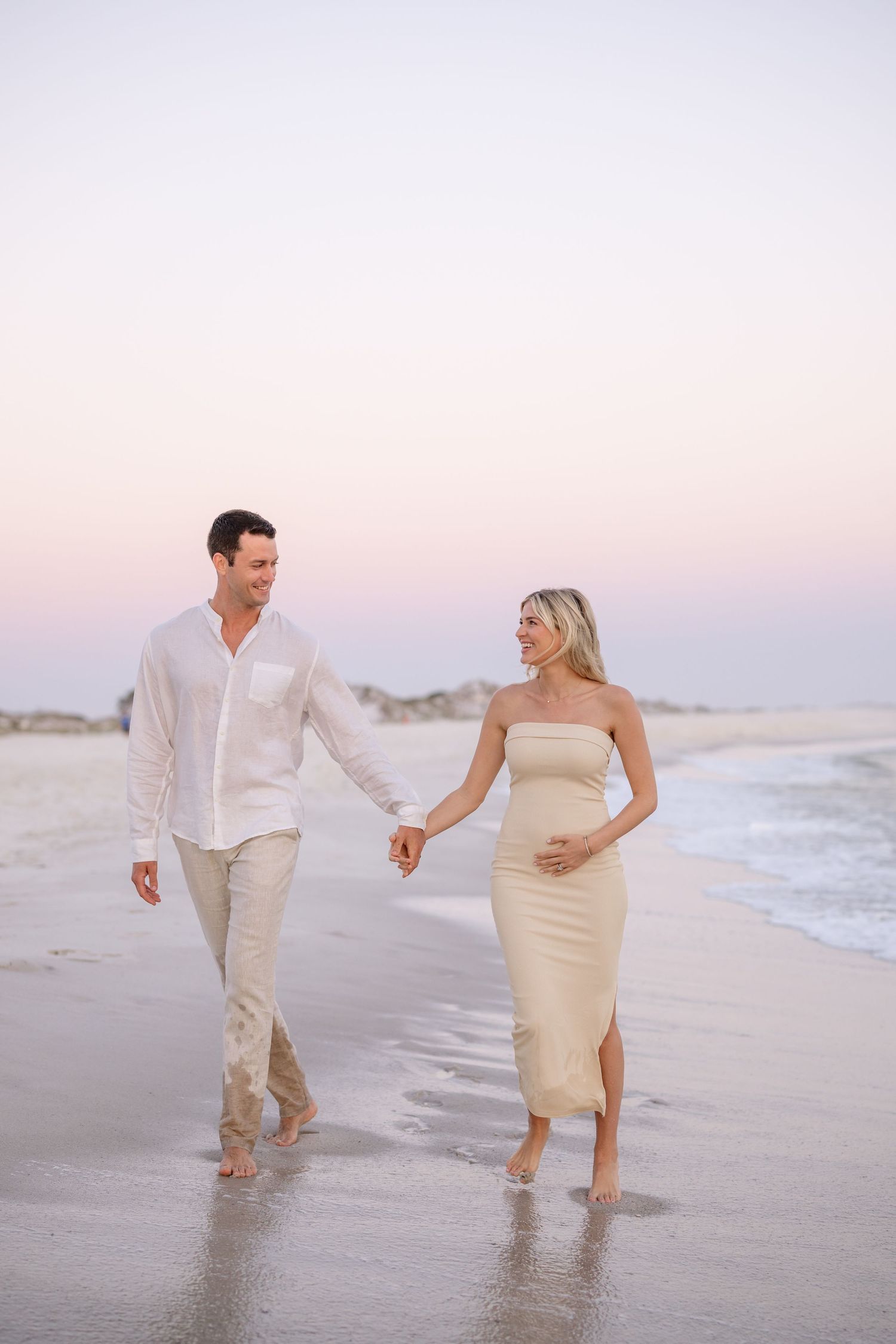 Couple walking barefoot along beach shore at sunset.