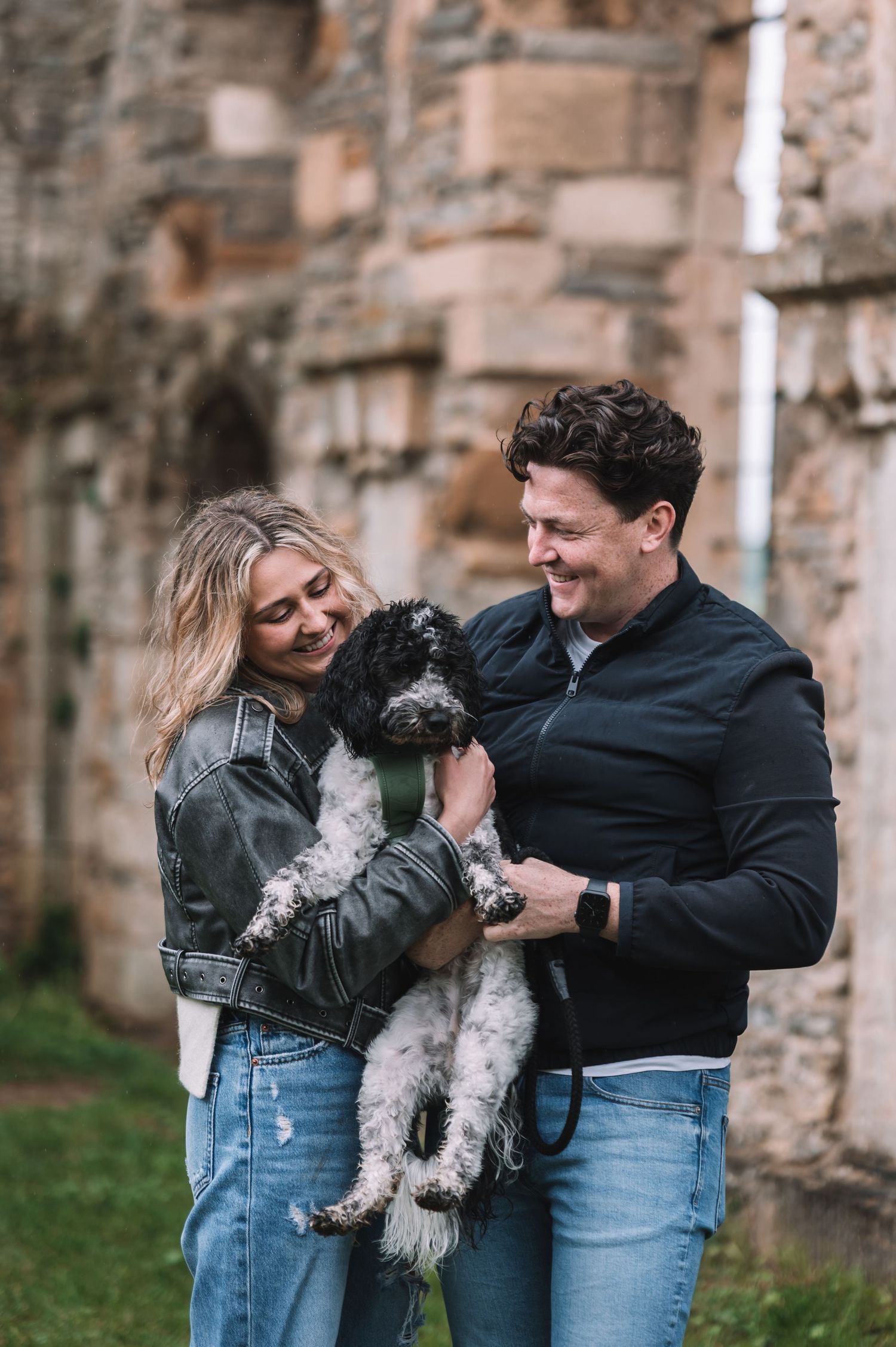 A couple in casual denim outfits joyfully plays with their fluffy black and white dog against a stone wall backdrop.