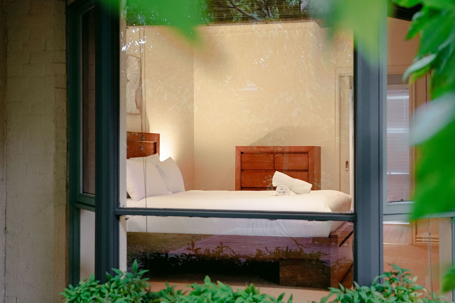 A cozy bedroom with wooden furniture viewed through an open doorway framed by green plants.