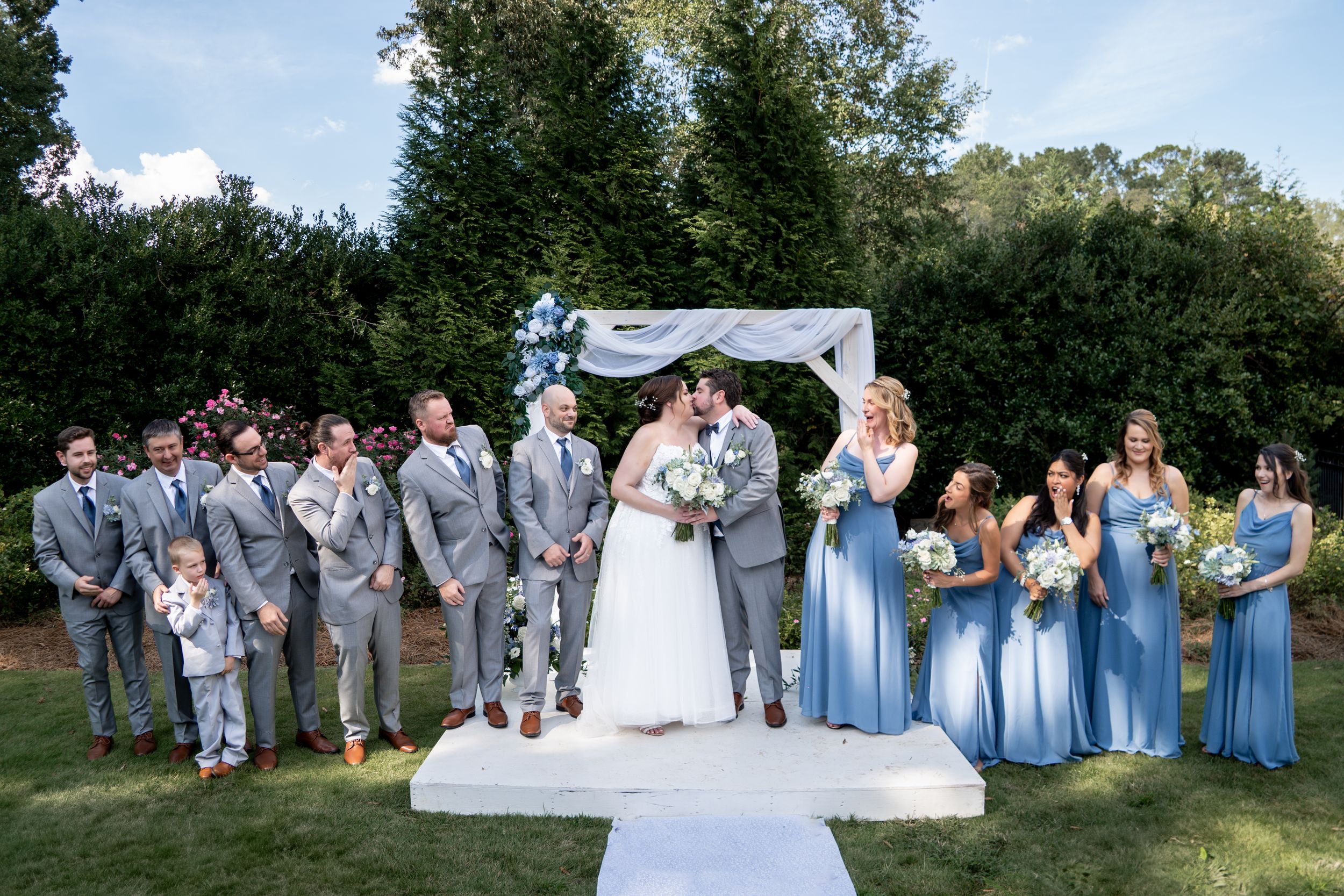 Wedding party in light blue and gray attire stands on white platform during outdoor garden ceremony.