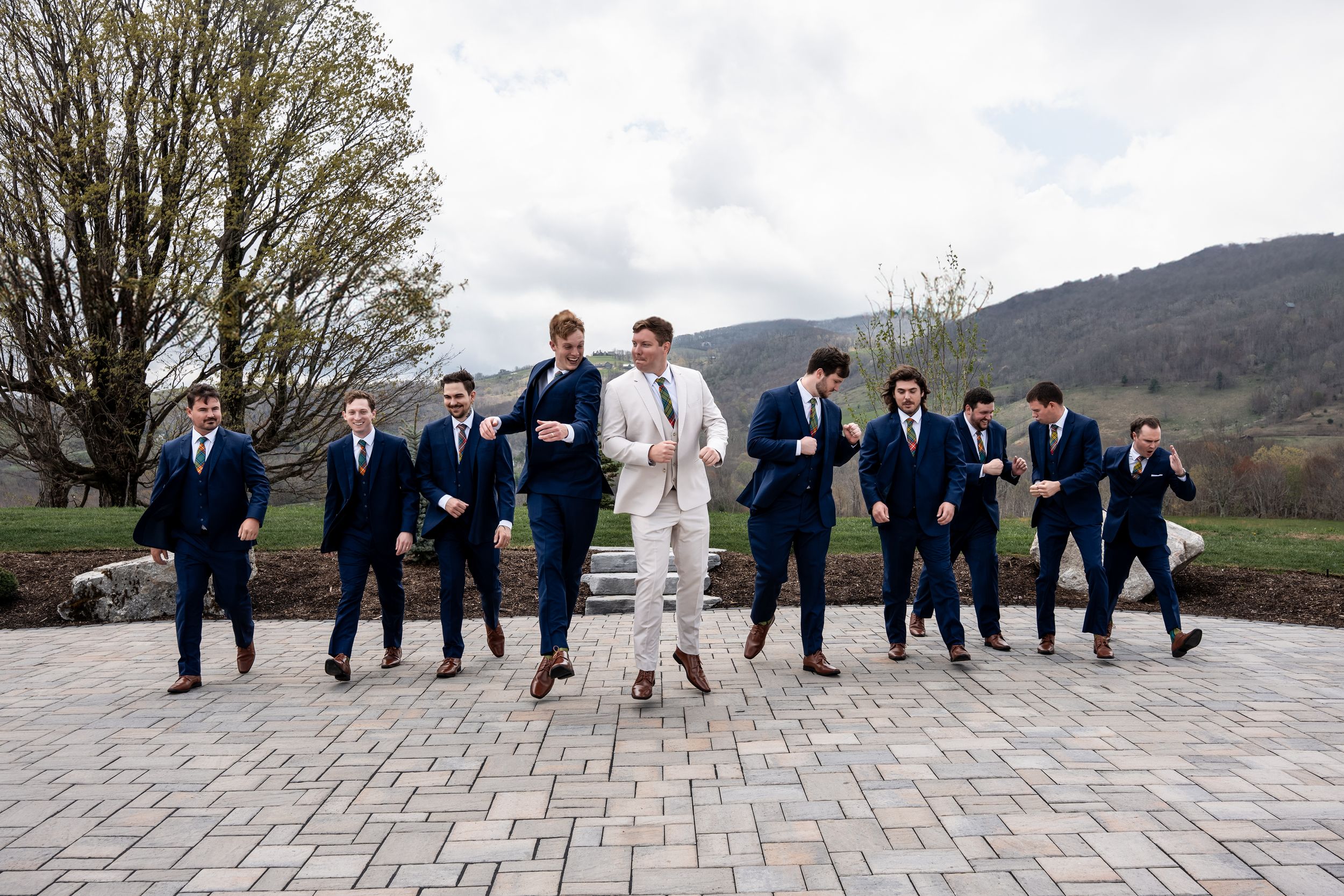 Wedding party in coordinated navy suits and one light colored suit jump in unison on stone patio with mountain backdrop.
