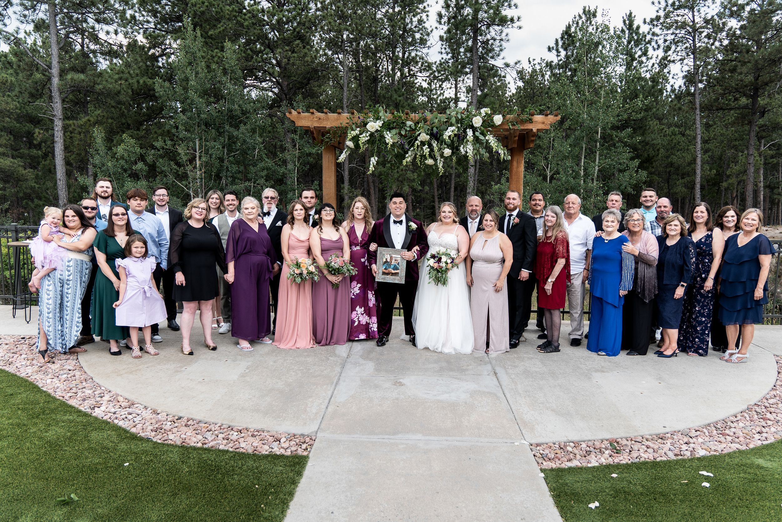 A wedding party poses outdoors on a curved stone patio with wooden pergola structures and pine trees in the background.